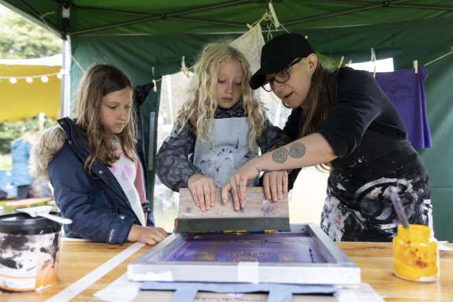 Workshop zeefdrukken onder een tent, twee meisjes leren zeefdrukken van een mevrouw met een pet op